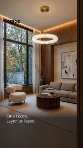 Modern living room with large windows, a beige sofa, round wooden coffee table, abstract artwork, and elegant chandelier.