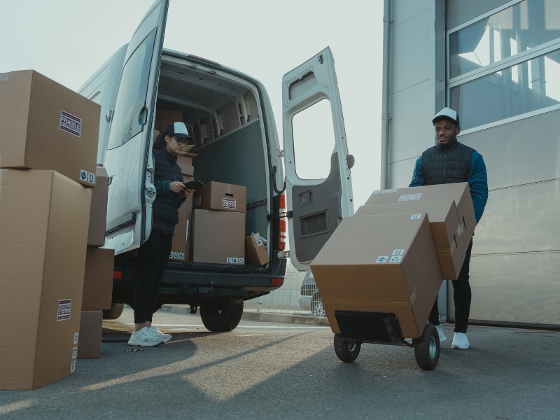 Delivery workers loading and unloading boxes from a van at a shipping facility, showcasing logistics and transportation operations.