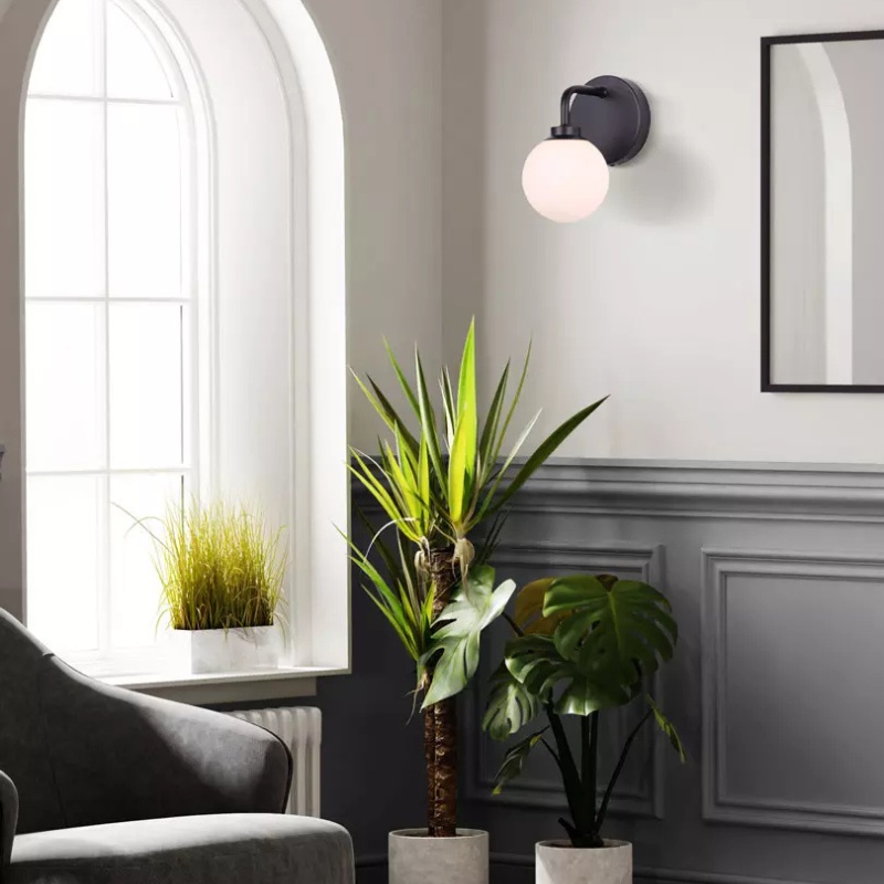Modern living room featuring a stylish wall sconce, two potted plants, and a cozy armchair, with natural light streaming through a large window.
