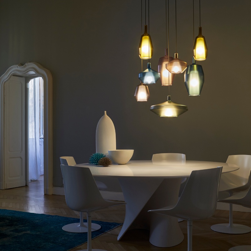 Modern dining room featuring a round white table surrounded by sleek chairs, illuminated by colorful pendant lights. A minimalist vase and decorative bowl add a stylish touch to the decor.