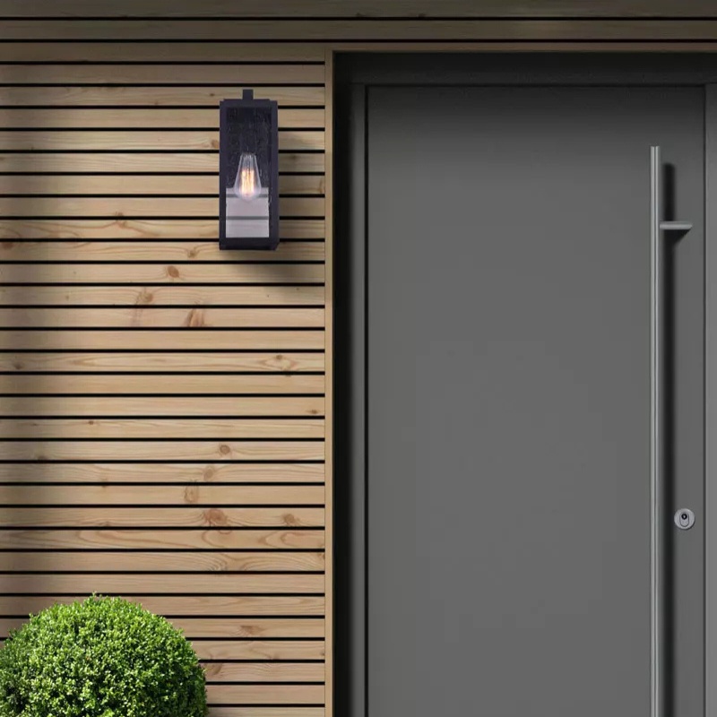Modern exterior entrance featuring a sleek dark wall lantern beside a gray door, complemented by a neatly trimmed green bush and wooden slat siding.